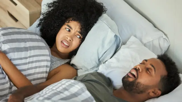 Mulher acordada na cama ao lado de parceiro que ronca, ilustrando quem percebe o ronco e as pausas na respiração